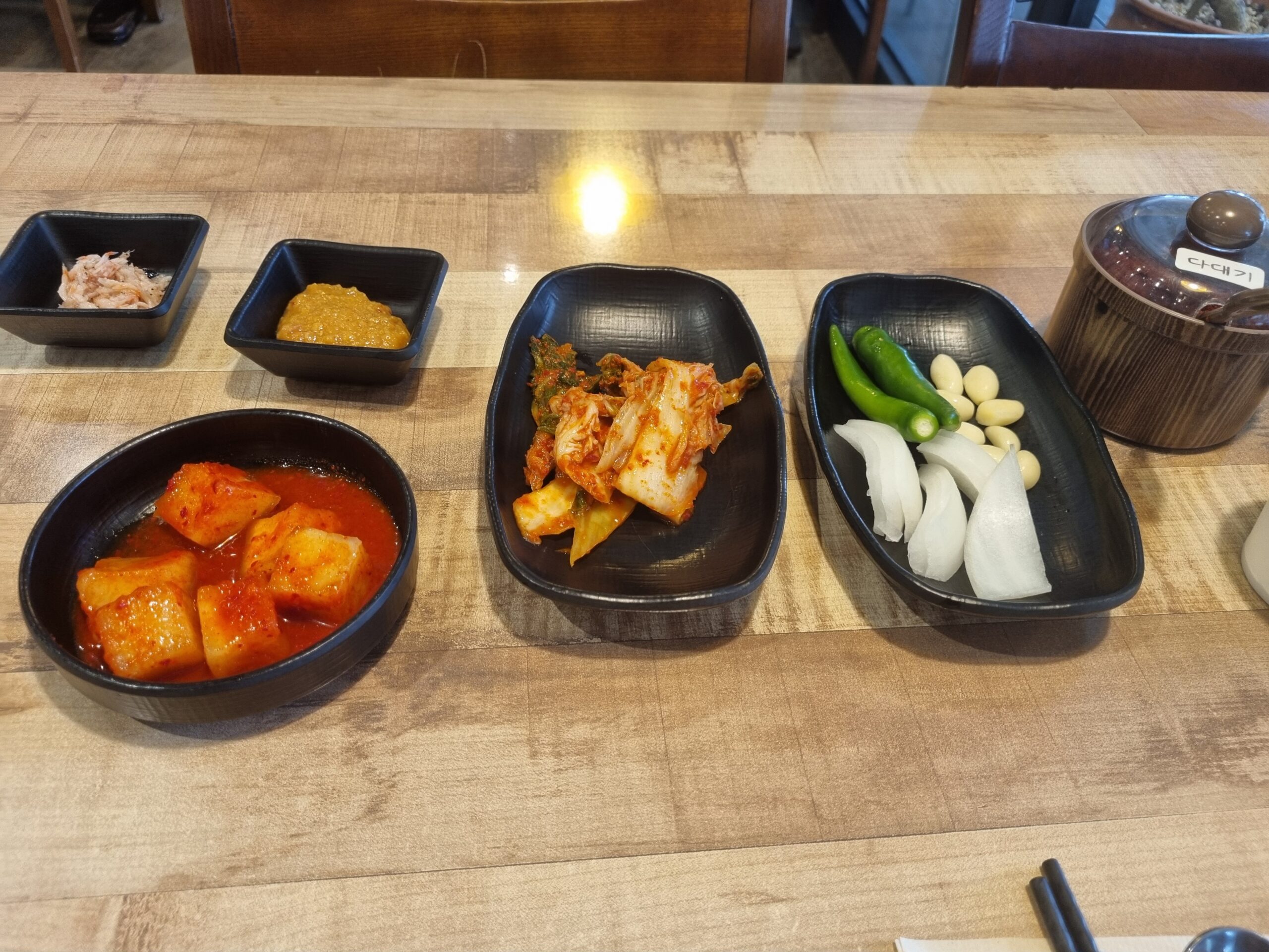 A tray of banchan at a Busan gukbap shop: kkakdugi, cabbage kimchi, pickled radish, raw garlic and chillies, with a pot of dadaegi