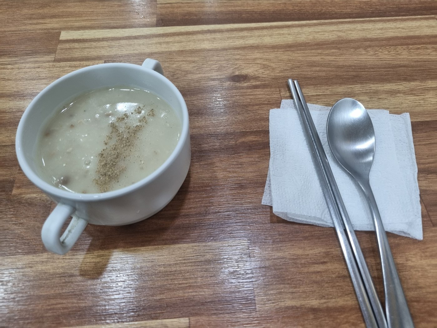 A cup of pale mushroom soup beside chopsticks and a spoon on a napkin