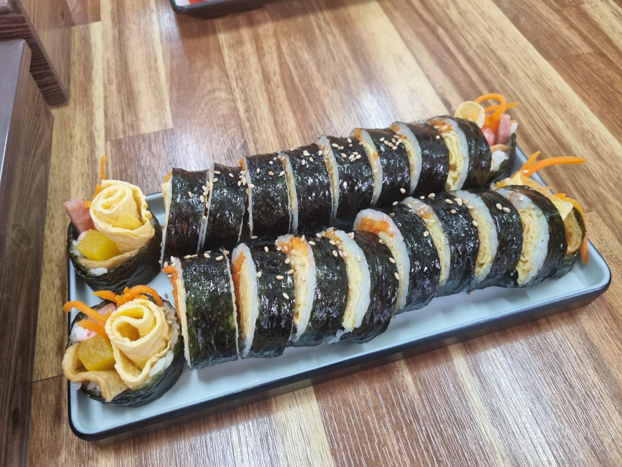 Gimbap on a wooden board with a side plate of pickled yellow radish and fresh kimchi